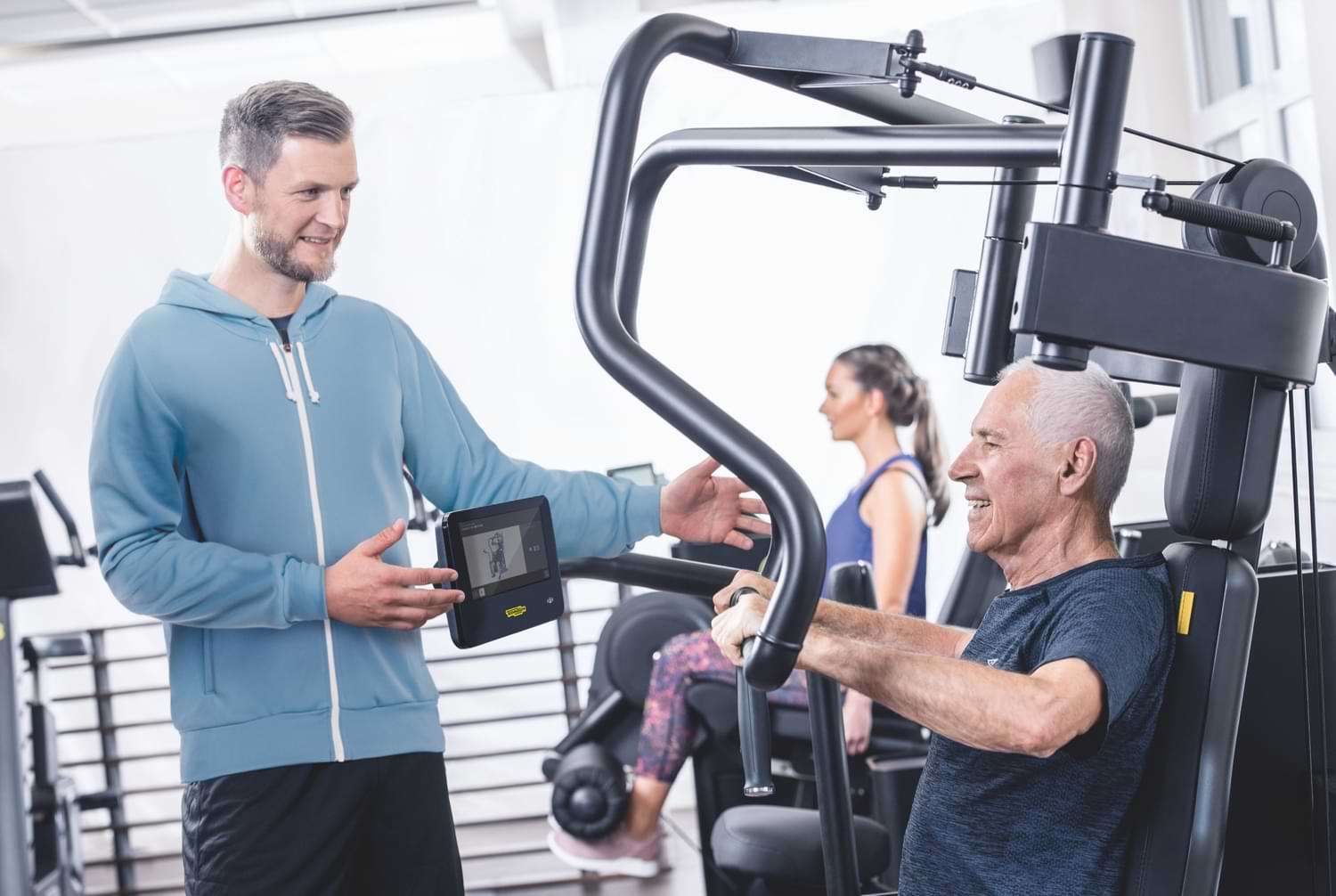 Ein Personal Trainer lächelt einen älteren Mann an der auf einem Sportgerät seinen Körper trainiert